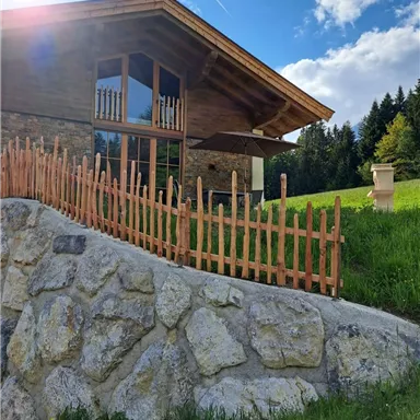 Ein schönes Holzhaus mit großen Fenstern und einem roten Zaun. Hinter dem Haus erstreckt sich eine grüne Wiese unter einem blauen Himmel.