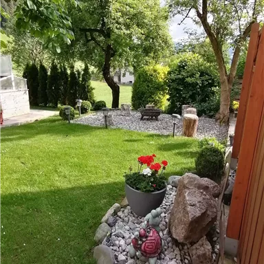 Ein schöner Garten mit bunten Blumen in einem Topf und einer kleinen Steinlandschaft. Im Hintergrund sind Bäume und eine grüne Wiese zu sehen.