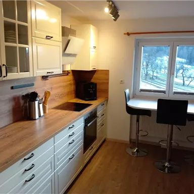 A modern kitchen with wooden surfaces and white cabinets. It features a dining table with tall chairs and large windows that let in daylight.