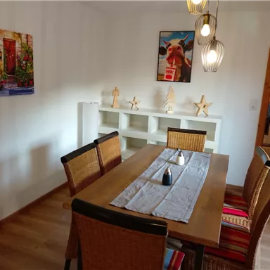 An inviting dining area with a wooden table and woven chairs. Two colorful pictures hang on the wall, while the table is adorned with a tablecloth and a decorative object.