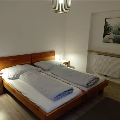 A leisurely bedroom with two wooden single beds and clear, modern furniture. Amenities include a radiator and a painted landscape picture on the wall.