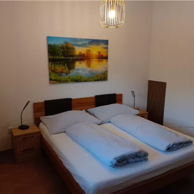 A cozy bedroom with a double bed and two pillows. On the wall hangs a picture of a landscape with a sunset.