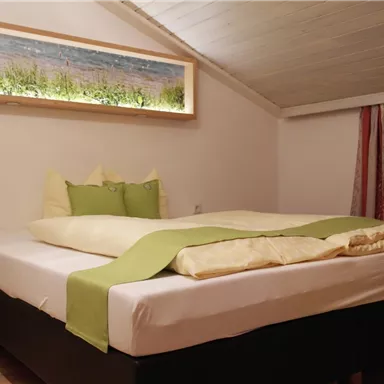 Ein gemütliches Schlafzimmer mit einem großen Bett und grüner Bettwäsche. Hinter dem Bett befindet sich ein beleuchtetes Wandpaneel mit einem schönen Landschaftsbild.