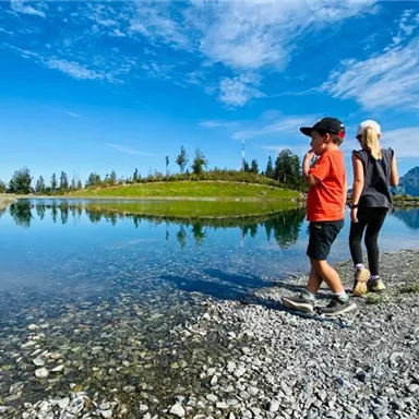 Two people walk along a clear lake, surrounded by greenery and mountains. The sky is blue with some clouds.