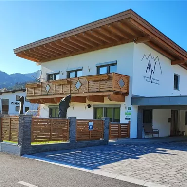 A modern guesthouse with wooden cladding and a large balcony. In the foreground, there is an entrance area with a hedge-like fencing.