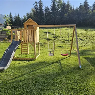 Ein Spielplatz mit einer Rutsche und zwei Schaukeln steht in einem grünen Garten. Im Hintergrund sind Bäume und ein hellblauer Himmel sichtbar.
