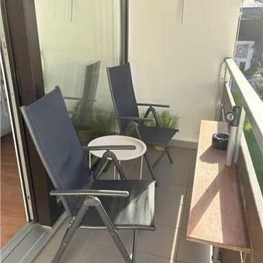 A relaxed balcony with two folding chairs and a small table. The sun is shining and there is a green plant in the background.