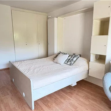 A bright room with a bed that is well organized. There is a shelf on the wall and a comfortable armchair in the corner.