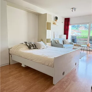 A cozy bedroom with an open bed and a comfortable armchair. Large windows provide plenty of daylight and a view of the surroundings.