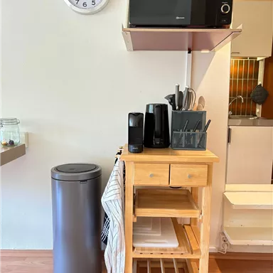 A compact kitchenette with a wooden table, microwave, and various kitchen appliances. Next to the table is a trash can and a wall with a clock.