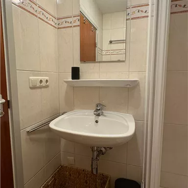 A small bathroom with a sink and a mirror. Bright tiles and a shower stall are visible on the wall.