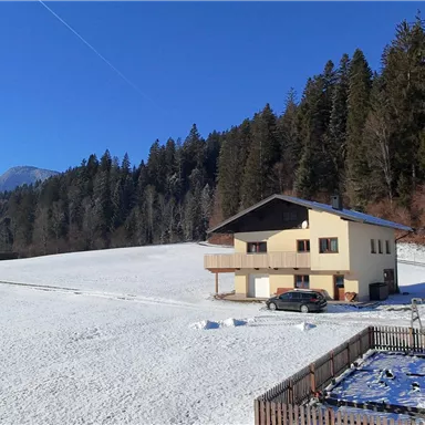 A cozy house stands in a snow-covered field, surrounded by trees. In the background, mountains and a clear blue sky are visible.