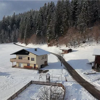 A snow-covered landscape with a residential house and adjacent buildings. In the background, tall trees and a clear sky can be seen.