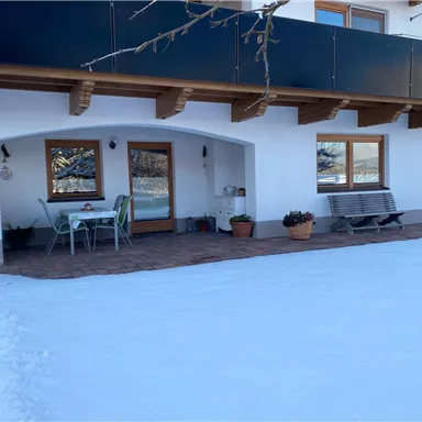 Ein gemütlicher Patio an einem Haus, umgeben von Schnee. Tische und Stühle laden zum Entspannen ein.