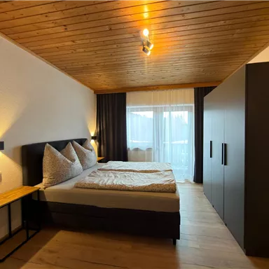 Ein modernes Schlafzimmer mit einem großen Bett und Holzdecke. Der Raum hat große Fenster und einen stilvollen Kleiderschrank.