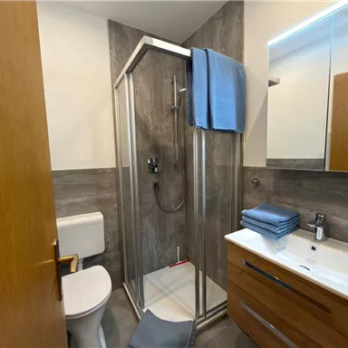 Ein modernes Badezimmer mit einer Dusche und Holzregalen. Die Wände sind mit grauen Fliesen verkleidet, und es gibt Handtücher und Pflegeprodukte auf dem Waschbecken.