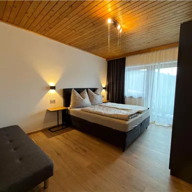 Ein gemütliches Schlafzimmer mit einem Doppelbett und zwei Kissen. Es gibt ein Sofa und große Fenster, die viel Tageslicht hereinlassen.