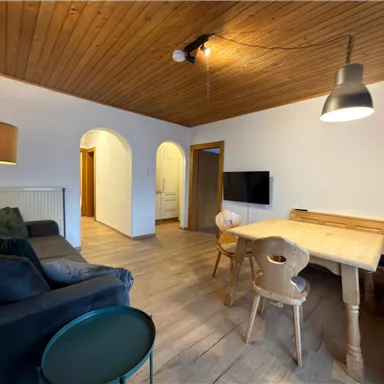 Ein gemütliches Wohnzimmer mit einem Sofa, einem Tisch und Stühlen. Das Zimmer hat eine helle Holzdecke und einen Fernseher an der Wand.