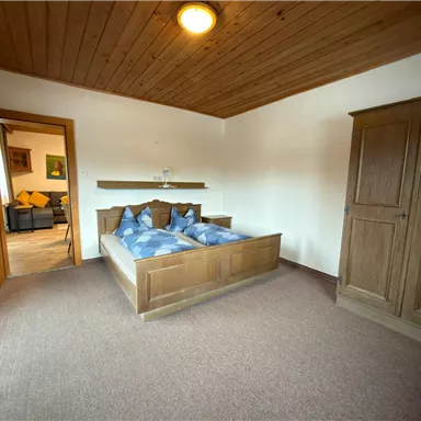 Ein gemütliches Schlafzimmer mit einem Doppelbett und blauen Kissen. Im Hintergrund ist ein Schrank und eine offene Tür zu einem weiteren Raum zu sehen.