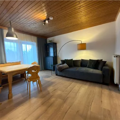 Ein gemütliches Wohnzimmer mit einem Holztisch, einer Couch und großen Fenstern. Die Einrichtung ist schlicht und modern, mit warmem Holz und sanften Farben.