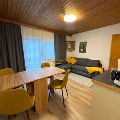 Ein gemütlicher Wohnraum mit einem Esstisch und gelben Stühlen. Im Hintergrund befindet sich eine Couch und eine Fensterfront, die natürliches Licht hereinlässt.