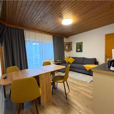 Ein gemütliches Wohnzimmer mit einem Essbereich und einem Sofa. Große Fenster sorgen für viel Tageslicht.