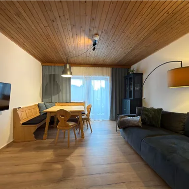 Ein modernes Wohnzimmer mit Holzdecke, gemütlichem Sofa und einem Essbereich. Große Fenster lassen viel Tageslicht herein.