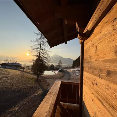 Ein Blick von einem Balkon auf eine malerische Landschaft bei Sonnenaufgang. Im Hintergrund sind Berge und Bäume zu sehen, während die Sonne den Himmel erleuchtet.