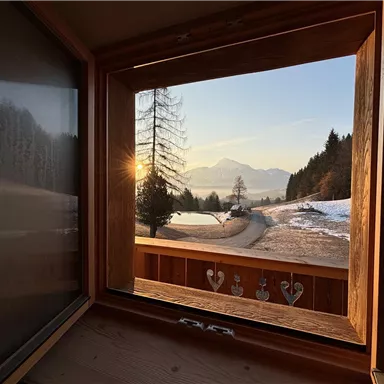 Ein wunderschöner Blick aus einem Fenster auf eine schneebedeckte Landschaft mit Bergen im Hintergrund. Die Sonne geht auf und taucht die Szene in warmes Licht.