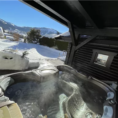 A hot tub in a snow-covered outdoor area with mountains in the background. The sun is shining, creating a relaxing atmosphere.