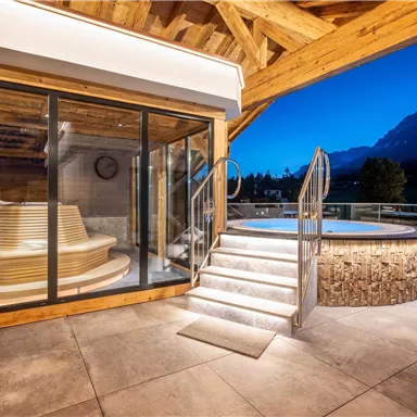 An elegant wellness area with a sauna and an outdoor whirlpool. The surroundings are surrounded by mountains and a clear evening sky.