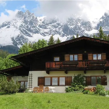 Ein hübsches alpenländisches Haus mit Holzbalkon, umgeben von grünen Wiesen. Im Hintergrund erheben sich majestätische, schneebedeckte Berge unter einem klaren Himmel.