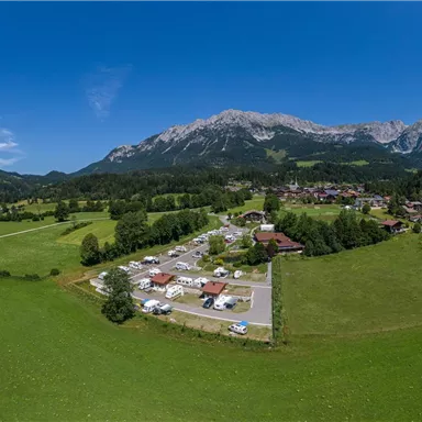 A vast, green landscape with mountains in the background. In the center, motorhomes and a small village are visible.