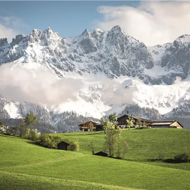 Ein wunderschönes Bergpanorama mit schneebedeckten Gipfeln und schönen Wolken. Im Vordergrund erstrecken sich saftige grüne Wiesen und eine malerische Landschaft.