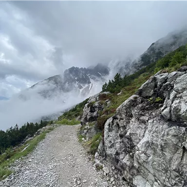 Ein schmaler Weg führt durch eine Gebirgslandschaft mit Felsen und dichter Nebel. Die Berge sind teilweise von Wolken umhüllt.