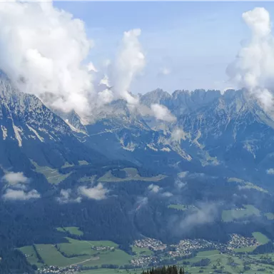 An impressive mountain landscape with high peaks and white clouds. In the foreground, green meadows and small settlements can be seen.