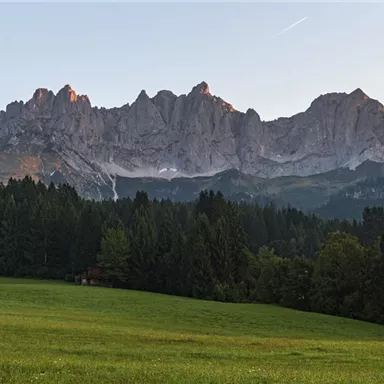An impressive mountain landscape with high rocks in the background and a green field in the foreground. The sun gently illuminates the peaks at dusk.