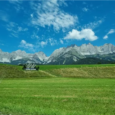 Eine grüne Wiese mit einem großen Schild im Vordergrund und majestätischen Bergen im Hintergrund. Der Himmel ist klar mit einigen Wolken.