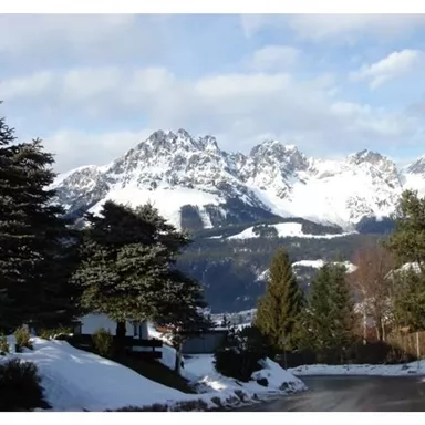 Eine schneebedeckte Berglandschaft mit hohen Gipfeln und grünen Tannenbäumen im Vordergrund. Der Himmel ist teilweise bewölkt, was eine ruhige Winteratmosphäre schafft.