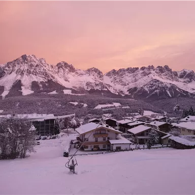 Eine malerische Winterlandschaft mit schneebedeckten Bergen und einem rosa Himmel. Im Vordergrund sind gemütliche Häuser in einer verschneiten Umgebung zu sehen.