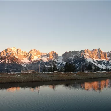 An impressive mountain landscape with snow-capped peaks and clear water in the foreground. The sky is blue and the sun's rays illuminate the mountains.