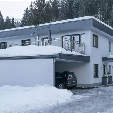 Ein modernes Haus im Schnee mit einer überdachten Terrasse. Im Vordergrund steht ein Auto vor der Garageneinfahrt.
