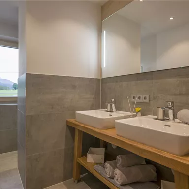 Ein modernes Badezimmer mit zwei Waschbecken und Holzregalen. Große Fenster sorgen für viel Tageslicht und einen Blick ins Grüne.