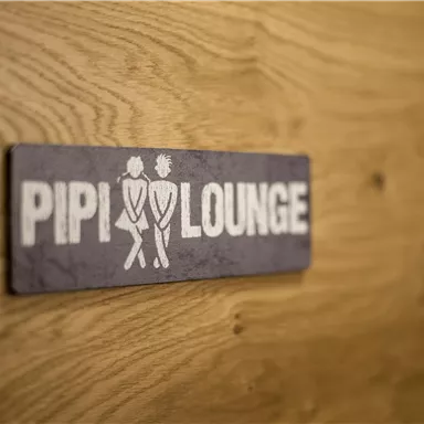 Ein Schild mit der Aufschrift "PIPI LOUNGE" auf einer Holzoberfläche. Es zeigt zwei stilisierte Figuren, die zusammen dargestellt sind.