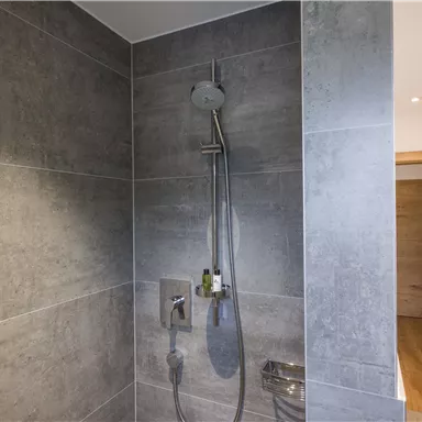 Eine moderne Dusche mit grauen Fliesen und einem schlichten Duschkopf. Im Hintergrund ist eine Holztür sichtbar.