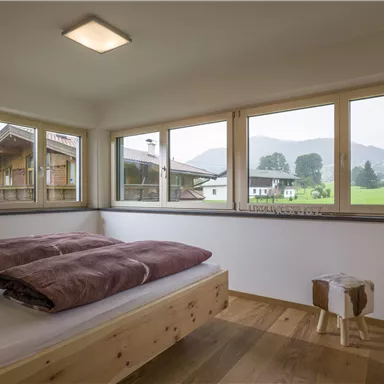 Ein gemütliches Schlafzimmer mit einem Holzbett und großen Fenstern. Durch die Fenster ist eine grüne Landschaft und die Berge im Hintergrund zu sehen.