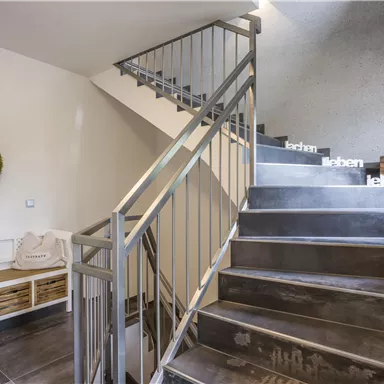 Ein moderner Treppenaufgang mit einem Geländer aus Metall. Auf der linken Seite befindet sich eine Sitzgelegenheit und eine grüne Wanddekoration.