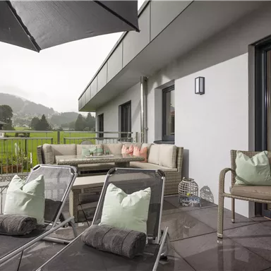 Eine moderne Terrasse mit bequemen Liegen und einem Sofa. Im Hintergrund sind grüne Wiesen und bewölkter Himmel zu sehen.