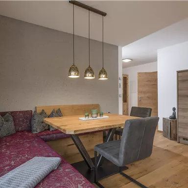 Ein gemütlicher Essbereich mit einer roten Sitzbank, einem Holztisch und modernen Stühlen. Die Beleuchtung wird durch drei hängende Lampen hervorgehoben.