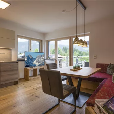 Ein modernes Wohnzimmer mit einer offenen Küche und großen Fenstern, die viel Licht hereinlassen. Ein gemütlicher Essbereich mit einem Tisch und einer Sitzbank lädt zum Verweilen ein.